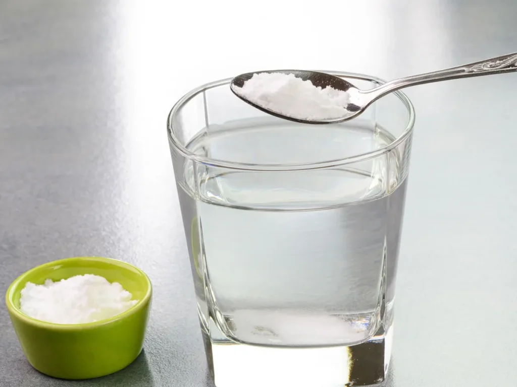 Um copo de água com uma colher cheia de Bicarbonato de Sódio em cima do copo, e na mesa também está um pequeno recipiente com Bicarbonato de Sódio.