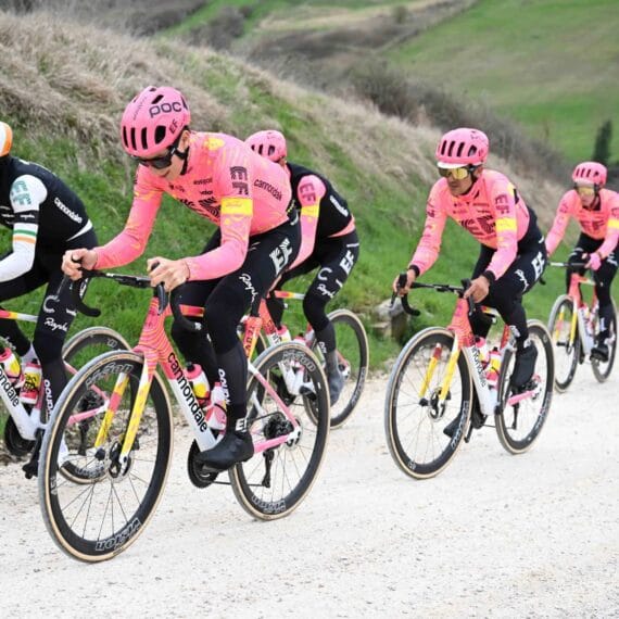 Cinco ciclistas profissionais treinando em uma estrada de montanha com o carro da sua equipe os seguindo.