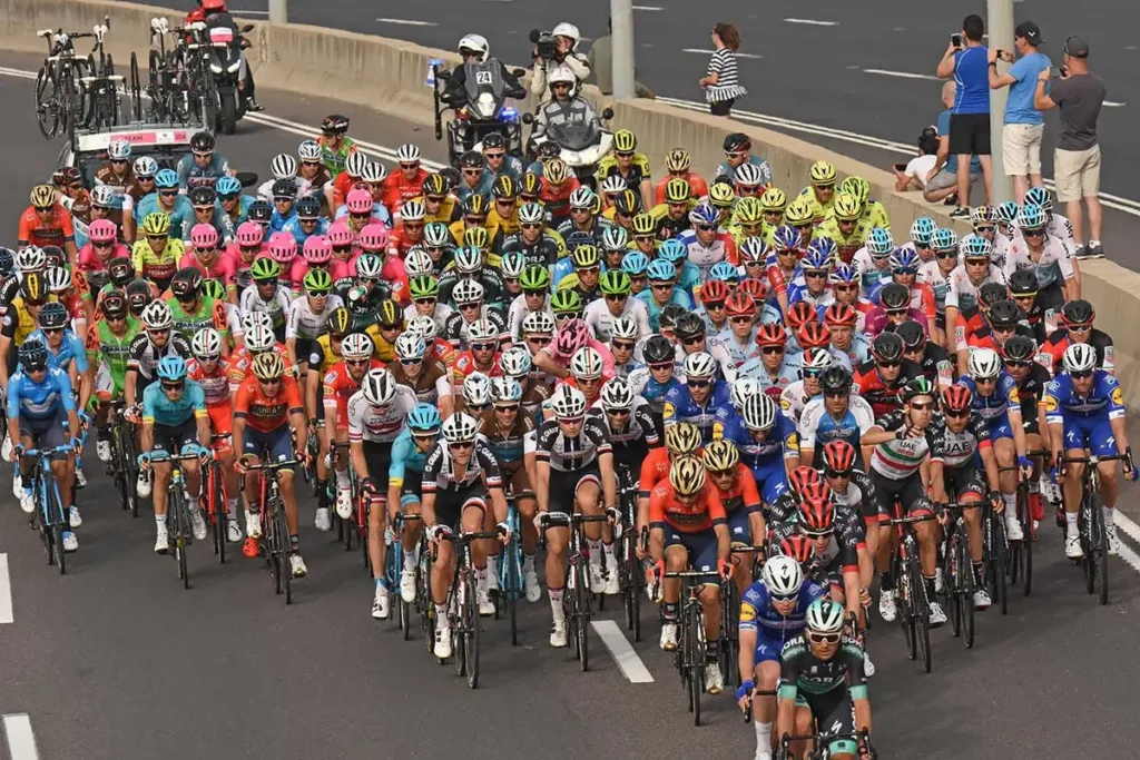 Ciclistas profissionais pedalando no formato de pelotão durante uma prova de ciclismo profissional.