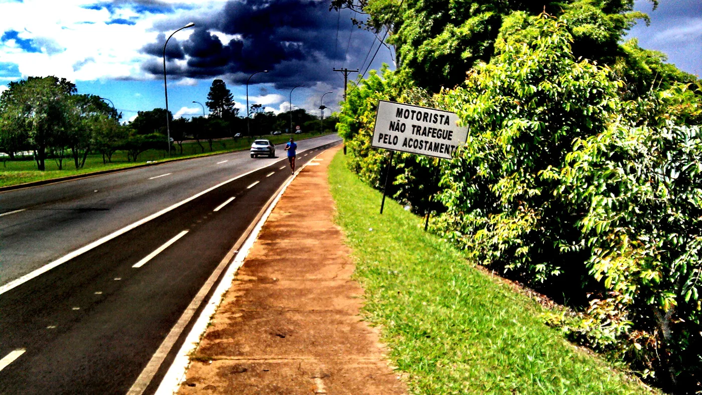 Acostamento de uma rodovia brasileira de pista simples, com placa "Motorista, não trafegue pelo acostamento" e um ciclista ao longe, no sentido do fluxo