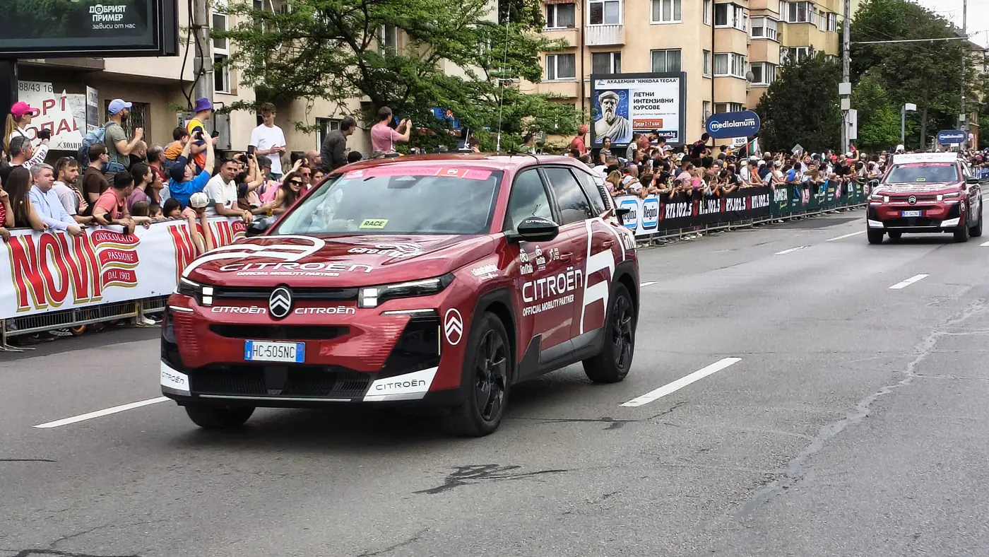 Carros oficiais do Giro d'Italia de 2026 numa avenida de Sófia, na Bulgária, com público atrás das grades e cartazes em cirílico ao fundo