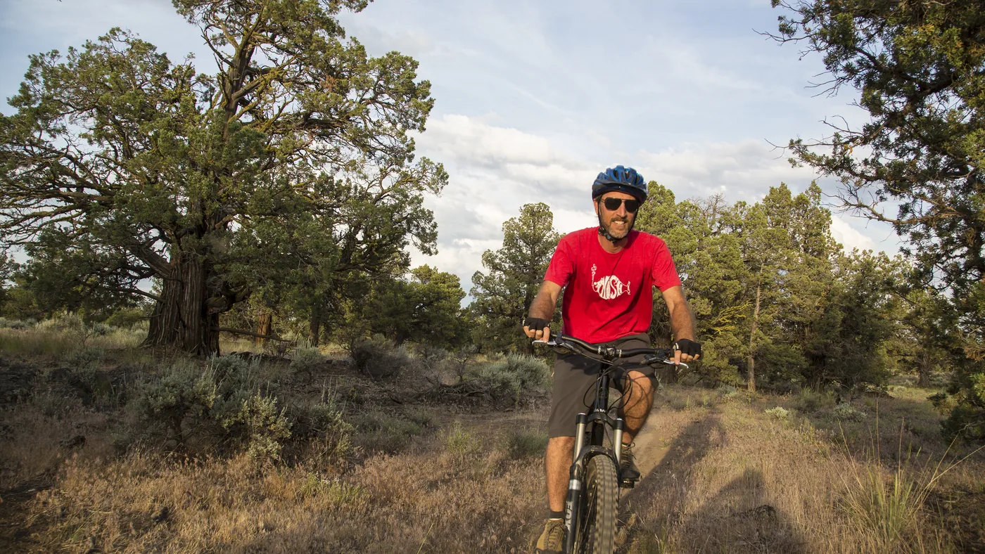 Ciclista pedala uma mountain bike numa trilha estreita de terra, entre arbustos e árvores baixas