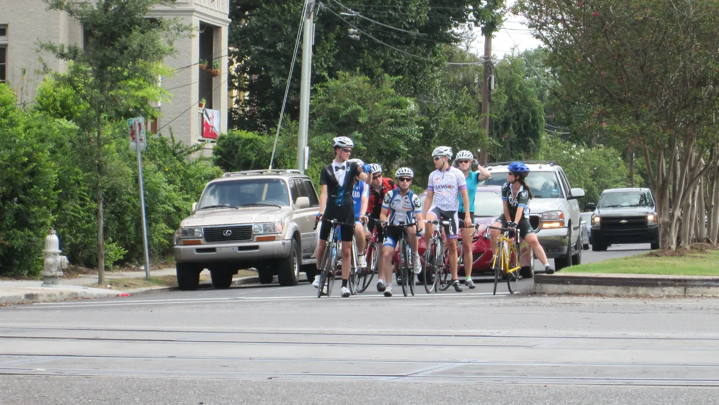 Grupo de ciclistas de estrada parados no sinal vermelho em um cruzamento urbano, com o pé no chão, à frente de carros que também aguardam