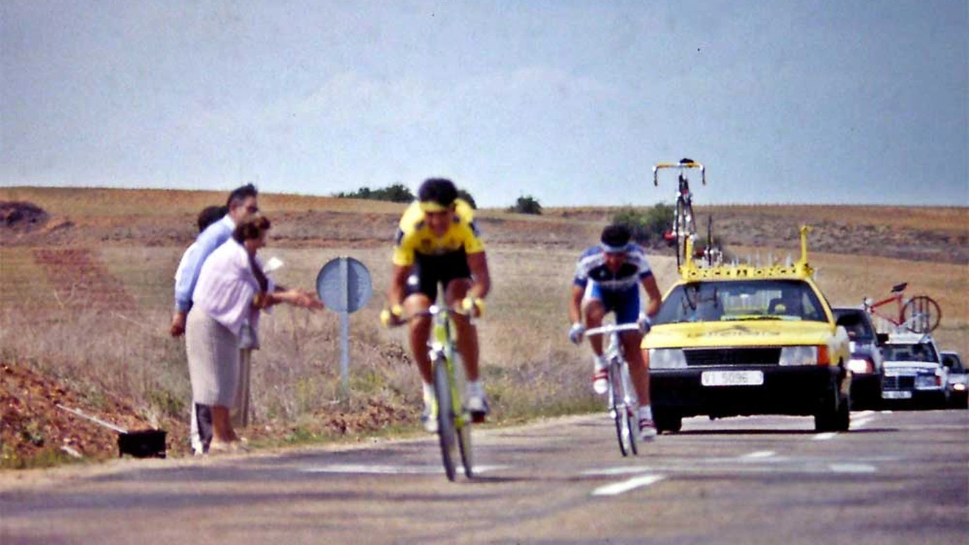 Dois ciclistas num contrarrelógio da Vuelta a España de 1989 numa estrada de Castela, o da frente de uniforme amarelo e preto, com carro de equipe e bicicletas reservas atrás
