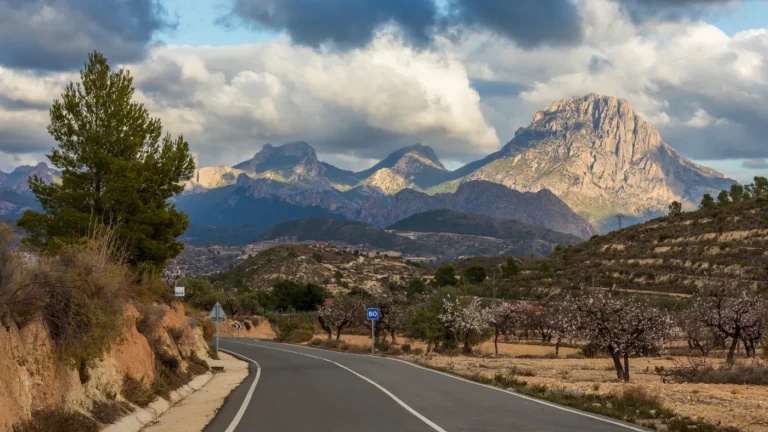 Estrada CV-775 vazia curvando em direção aos picos da Serra de Aitana, na província de Alicante, com amendoeiras em flor à direita e nuvens carregadas sobre a montanha