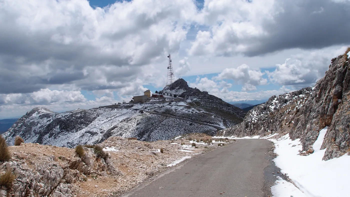 Estrada estreita subindo até o cume da Sierra de la Pandera, com a torre de antenas e as edificações da estação militar visíveis no alto e neve nas encostas