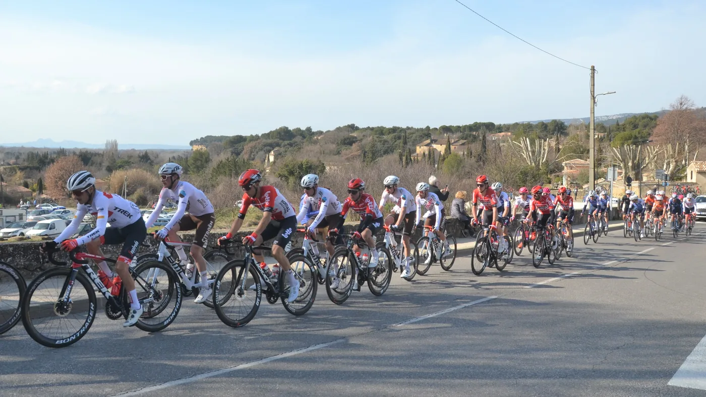Grupo de fuga do Tour de La Provence 2022 em fila numa estrada de campo com árvores e casas ao fundo, na etapa Arles > Manosque
