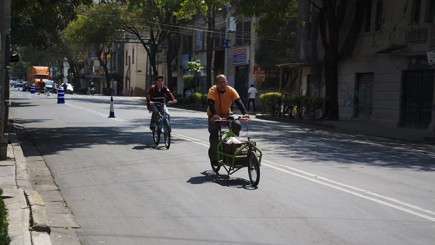 Ciclistas circulam pelo leito de uma rua urbana, um deles numa bike de carga de três rodas