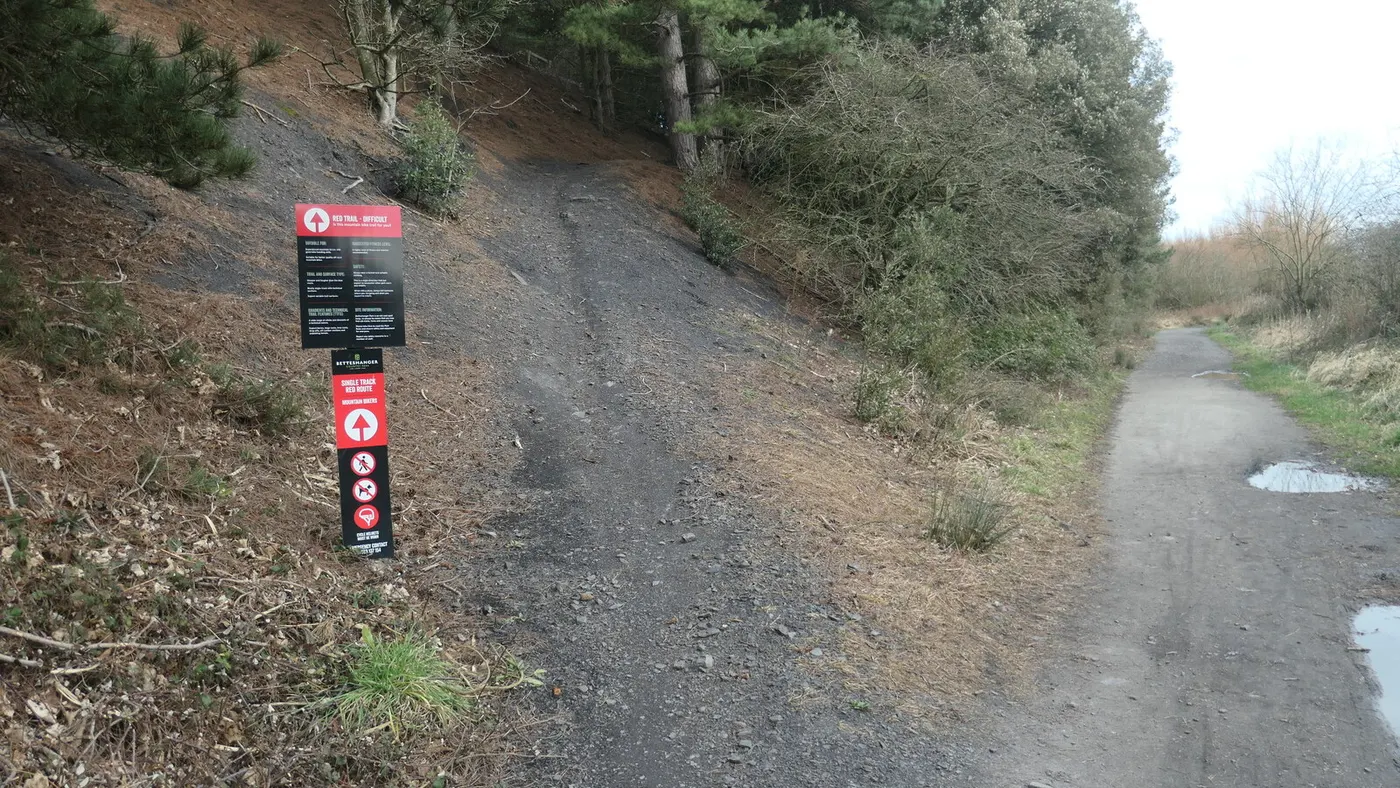 Entrada de trilha de mountain bike com subida de terra e placas de rota vermelha entre pinheiros