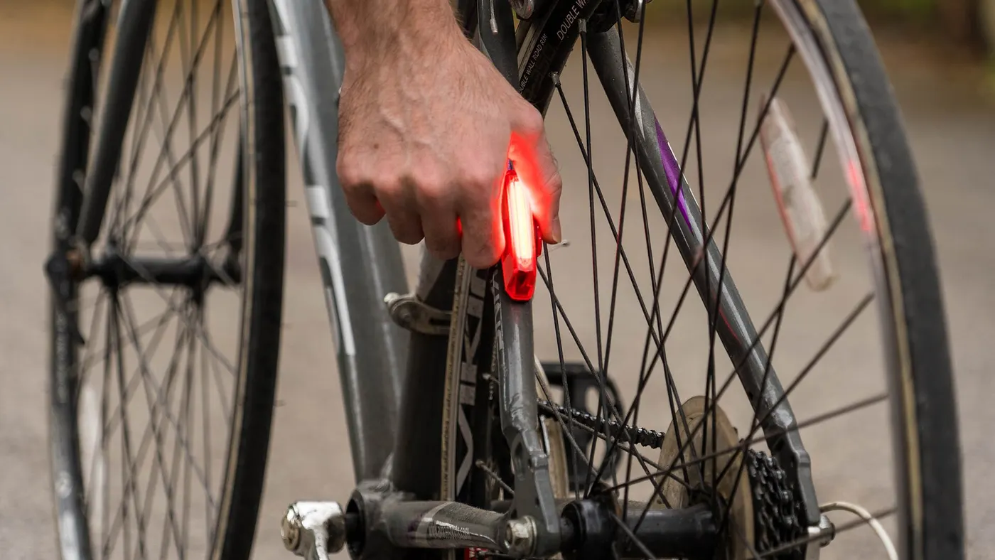 Mão aciona a luz traseira vermelha presa ao quadro de uma bicicleta, com refletor no pedal e na roda ao fundo